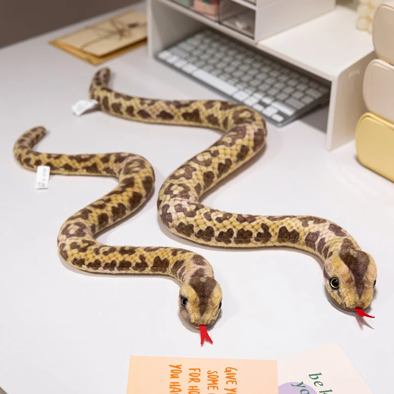 A realistic, soft plush toy of a patterned snake, likely an anaconda doll, lying on a white surface.
