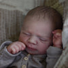 A close-up photo of a newborn baby doll wrapped in soft gray clothing, sleeping in a textured fabric setting.