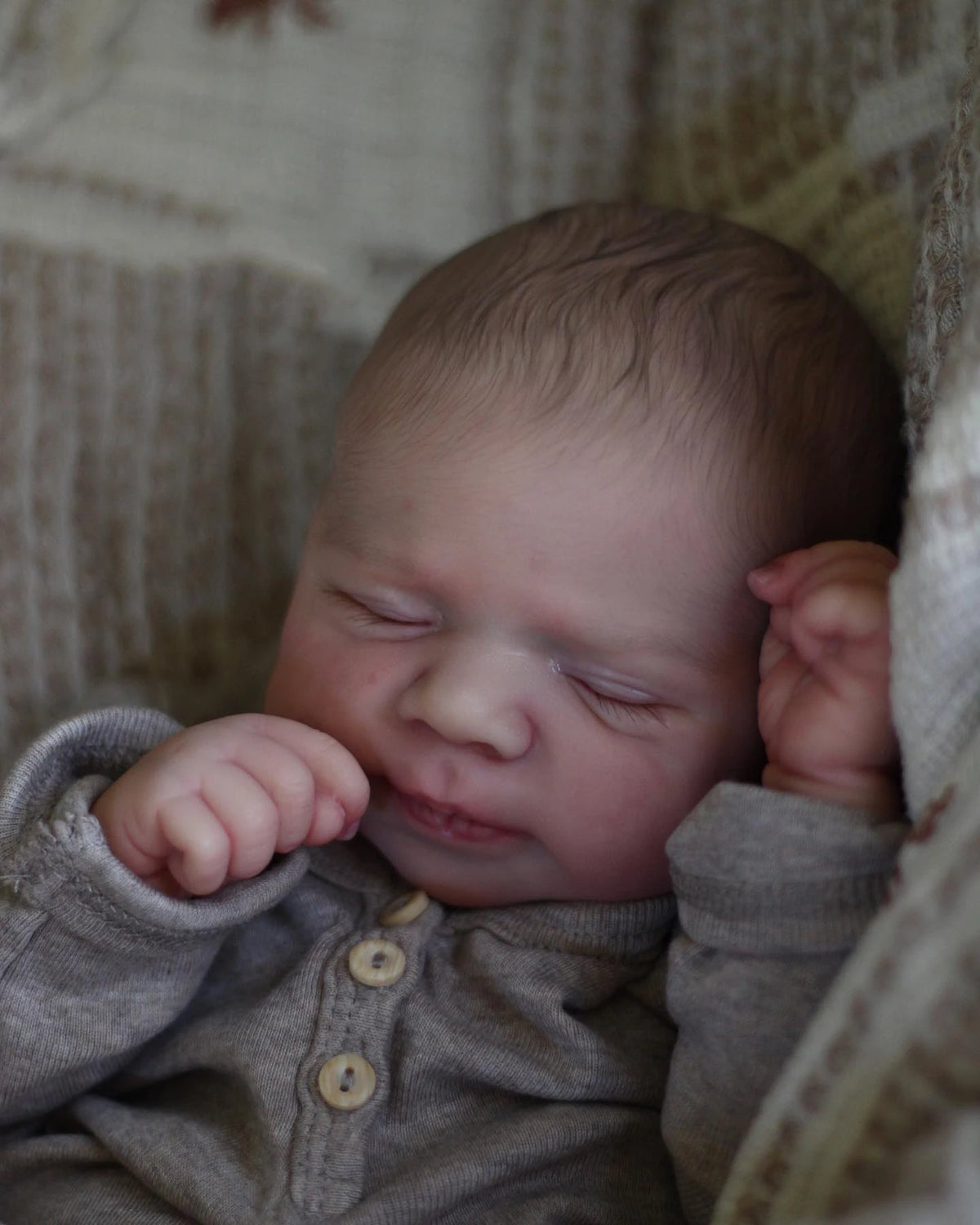 A close-up photo of a newborn baby doll wrapped in soft gray clothing, sleeping in a textured fabric setting.