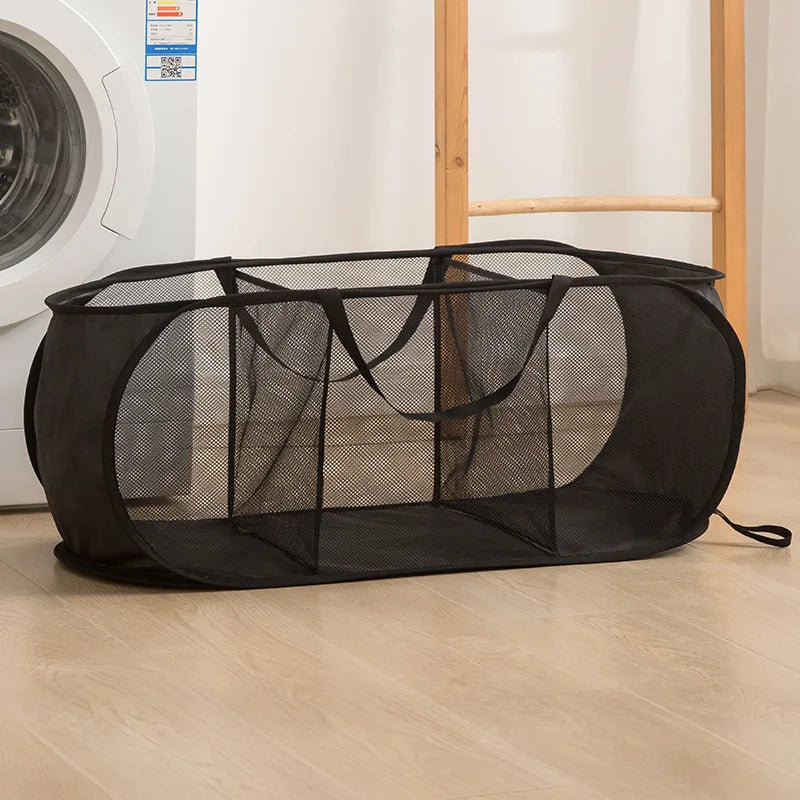 A black folding mesh laundry basket with handles resting on a light wooden floor.
