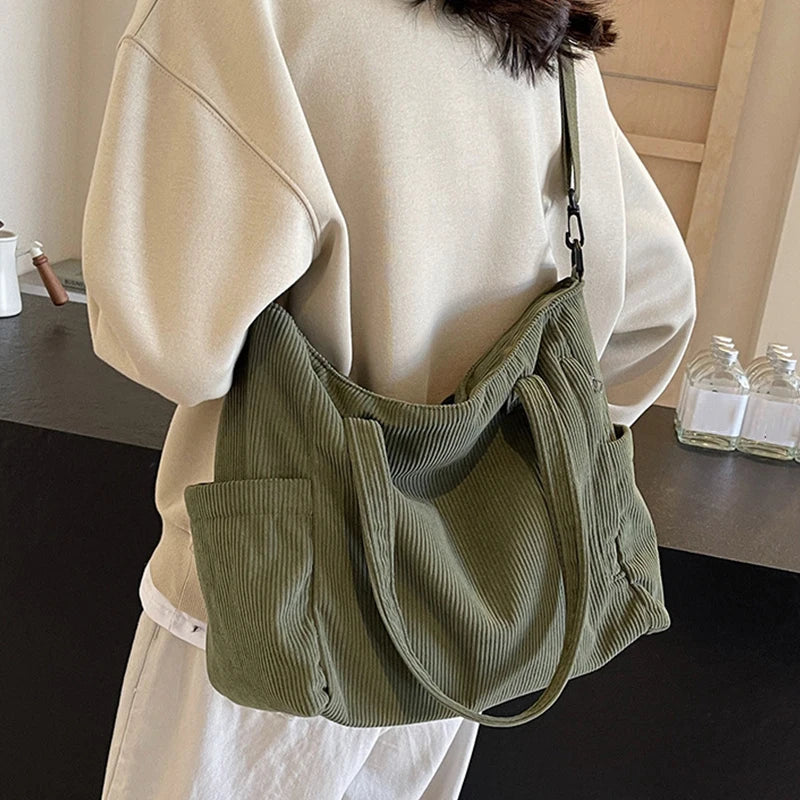 A light beige corduroy tote bag with a green strap is shown being carried by a person wearing a cream-colored jacket.