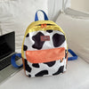 A yellow and white cow print backpack with blue straps resting on a laptop next to a white pillow.