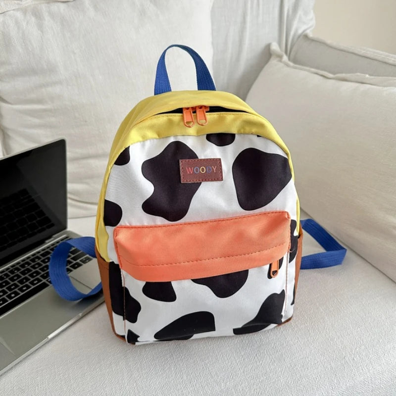 A yellow and white cow print backpack with blue straps resting on a laptop next to a white pillow.