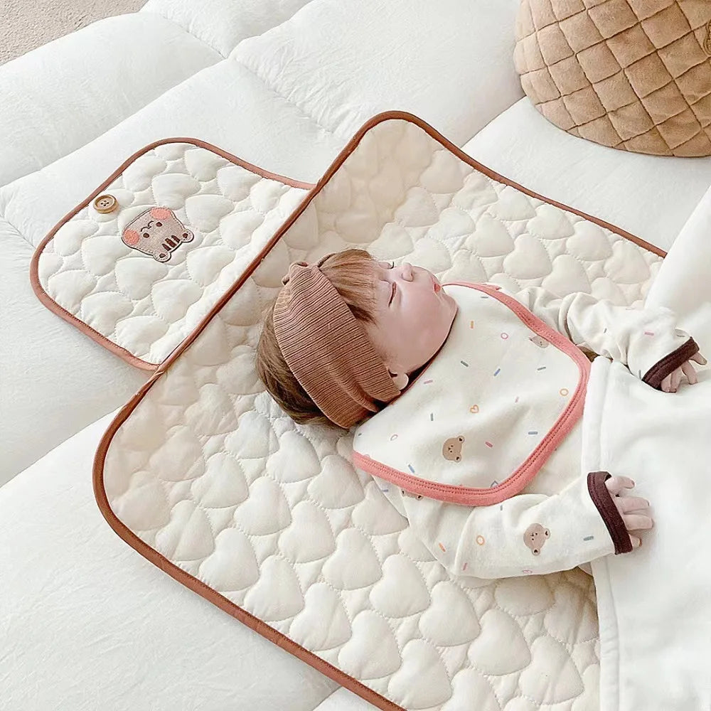 A baby is lying on a quilted beige and white portable diaper changing pad with a pink trim and matching clothing.
