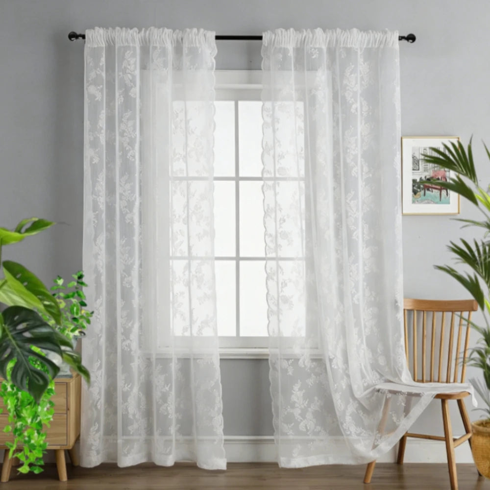 A pair of sheer white floral lace curtains hanging in a window with a white frame and light gray wall.