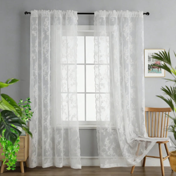 A pair of sheer white floral lace curtains hanging in a window with a white frame and light gray wall.