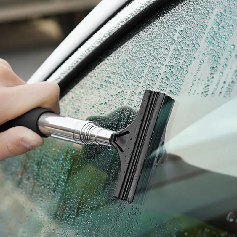 A hand uses a black retractable car mirror and window cleaning wiper to wipe water off a car window.