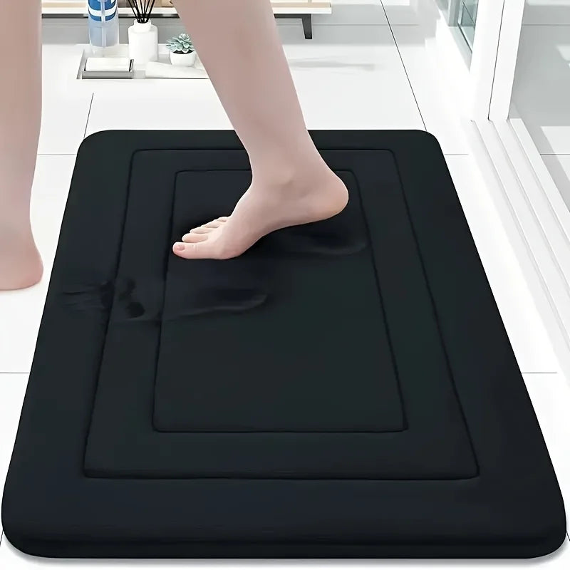 A black absorbent soft non-slip bath mat is placed on a white tiled floor, with a person's foot resting on it.
