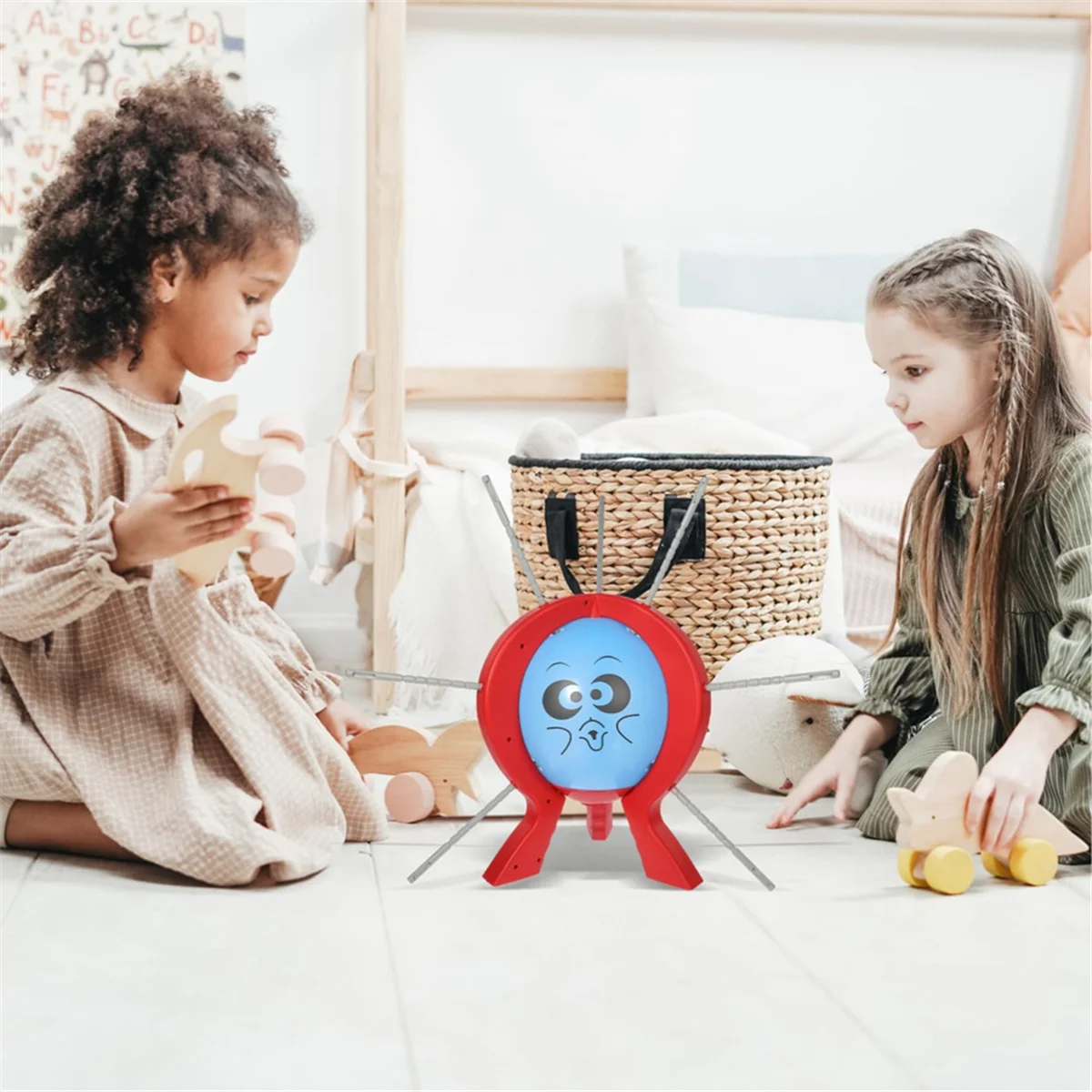 Two young girls play with a red balloon burst game on the floor in a bright room.