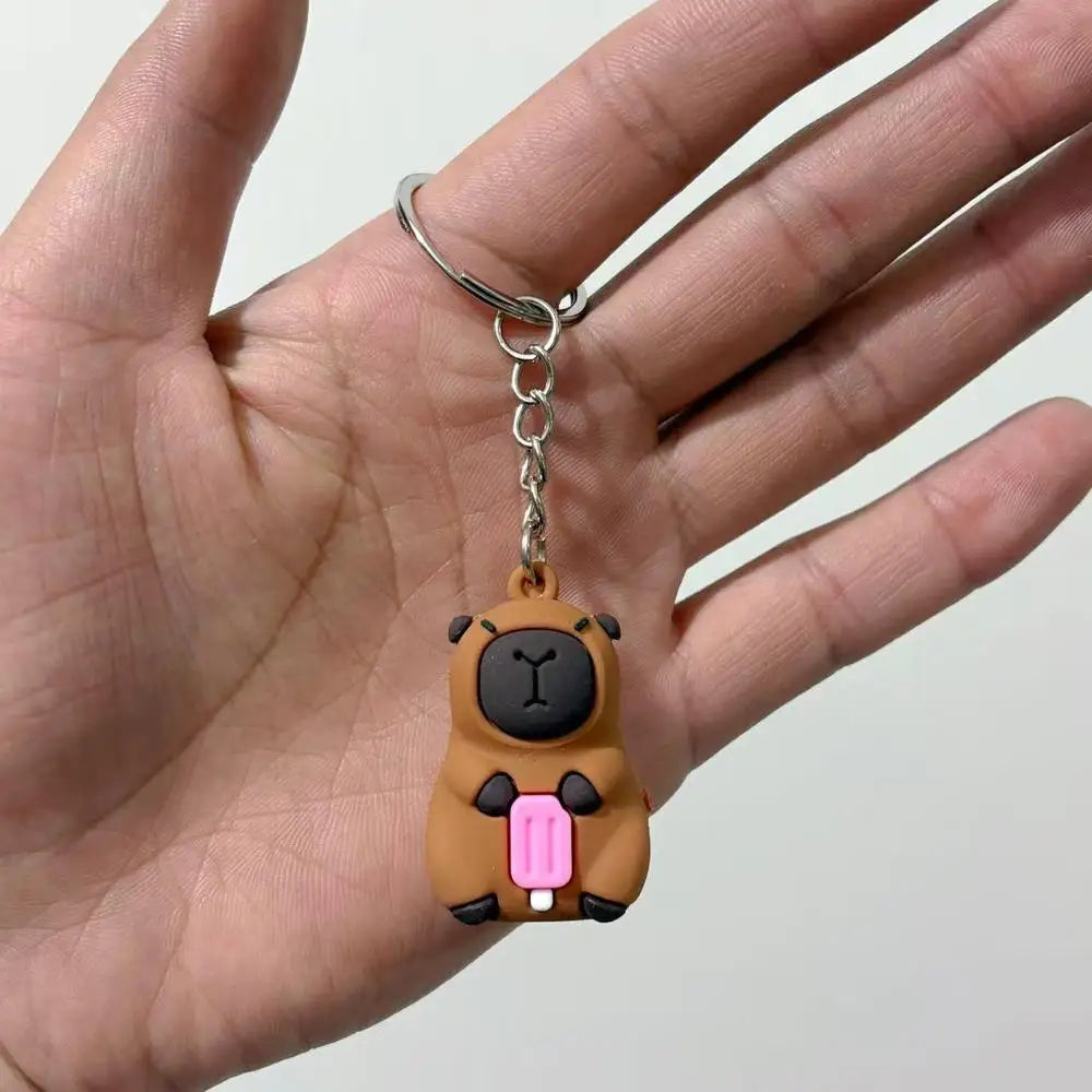 A small brown capybara charm with pink accents is being held in a hand, attached to a silver keyring.