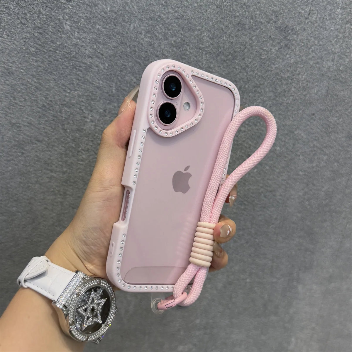 Una mano sostiene una funda de iPhone rosa con diseño de diamante y una correa de pulsera rosa.