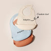 A set of three ultra-thin silicone baby bibs in cream, light blue, and terracotta, showing a custom text option.