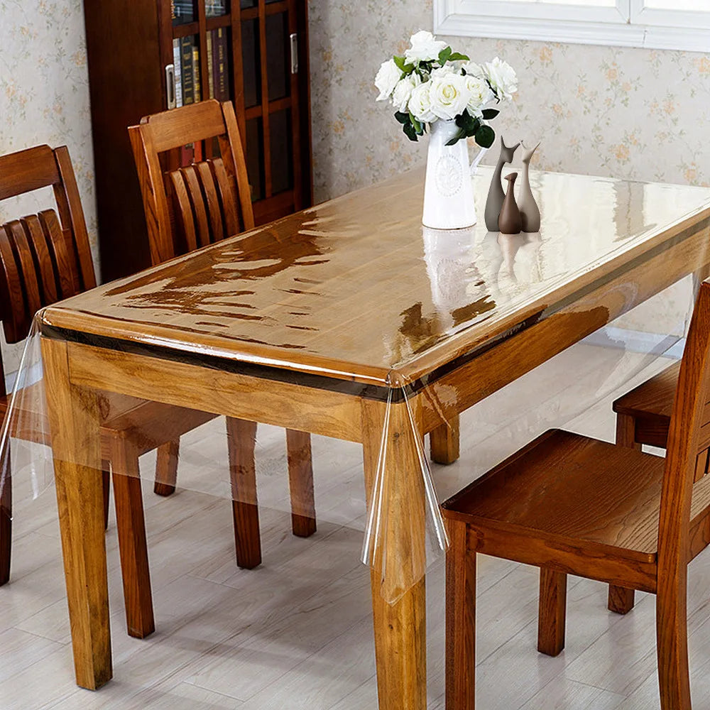 A wooden dining table with a clear PVC waterproof protector and wooden chairs, set in a bright room.
