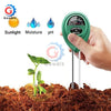 A hand holds a green soil testing meter with three probes inserted into dark soil, demonstrating light, moisture, and pH meas