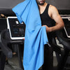 A man wearing a black tank top holds up a bright blue microfiber sports towel in a gym setting.