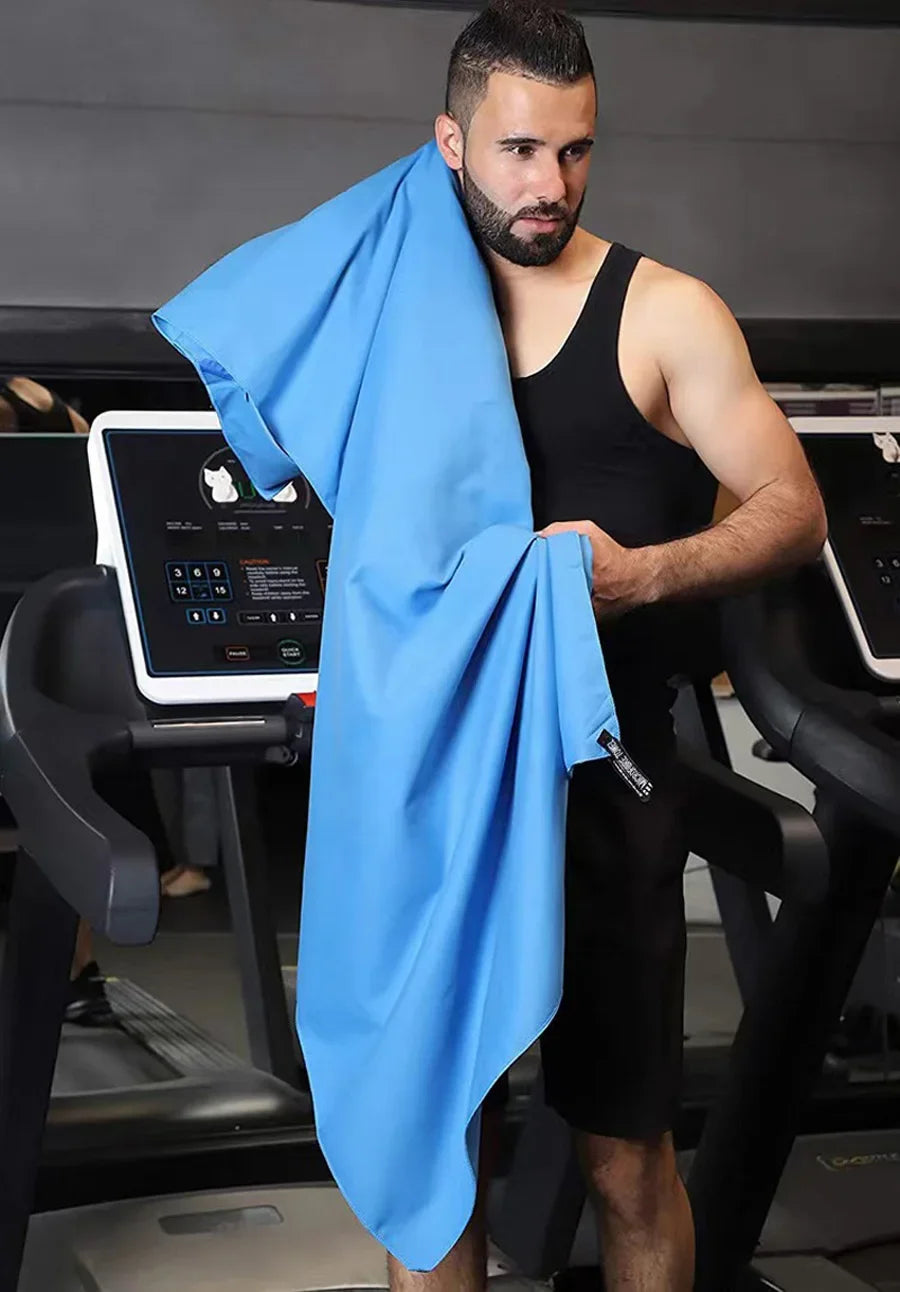A man wearing a black tank top holds up a bright blue microfiber sports towel in a gym setting.