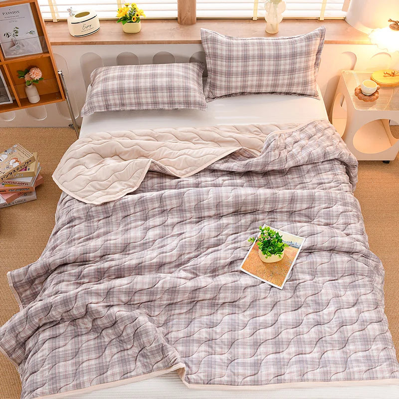 A plaid quilted blanket in light gray and white is displayed on a bed with pillows and a folded blanket.