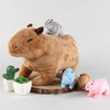 A brown stuffed capybara toy sits next to a gray and blue animal figurine and a green plant.