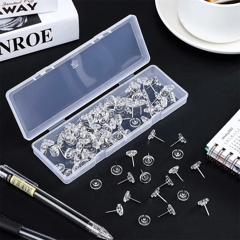 A transparent plastic box filled with clear thumb tacks and push pins is shown on a dark surface.