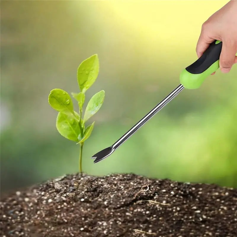 A hand holds a green ergonomic stainless steel weeder tool with soft-grip handles, positioned above soil and a small plant.
