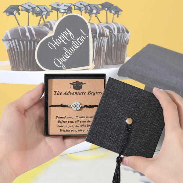 A hand holds an adjustable compass bracelet with a graduation cap box against a background of graduation-themed cupcakes.
