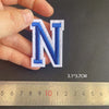 A hand holds a blue embroidered letter N patch, measured against a ruler.