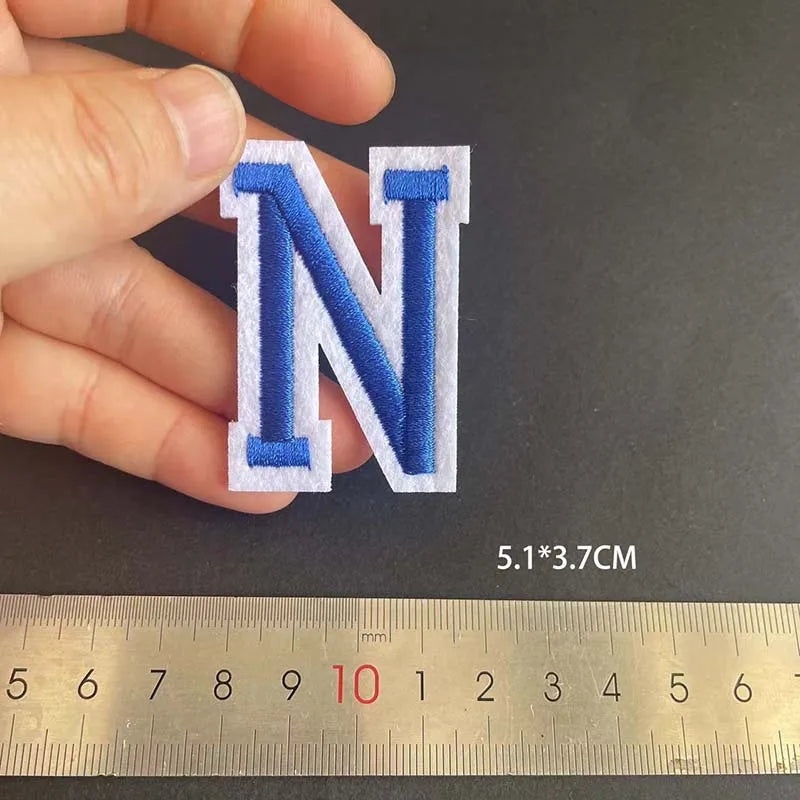 A hand holds a blue embroidered letter N patch, measured against a ruler.