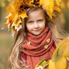 A young girl with long blonde hair wears a crown of yellow autumn leaves and an orange knitted scarf.