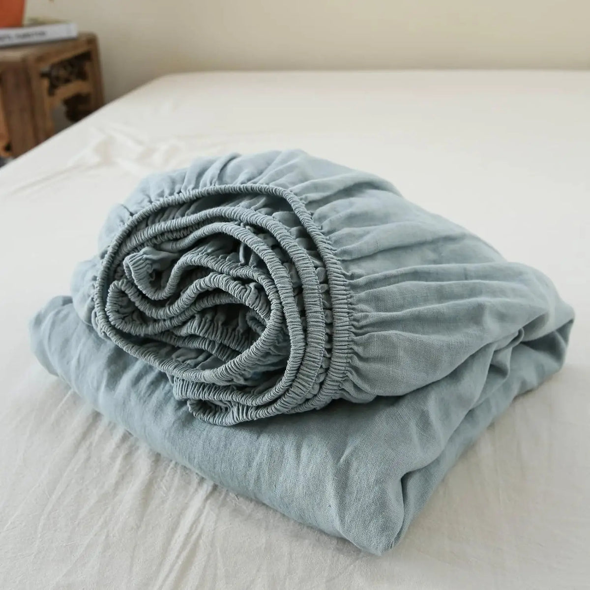 A tightly rolled light blue linen fitted sheet with deep pockets resting on a white bed.