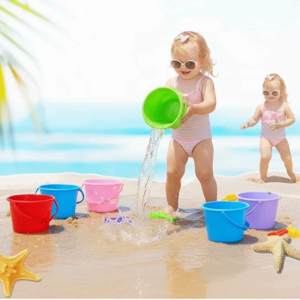 A toddler plays with a green plastic water bucket, surrounded by other colorful buckets on a sandy beach.