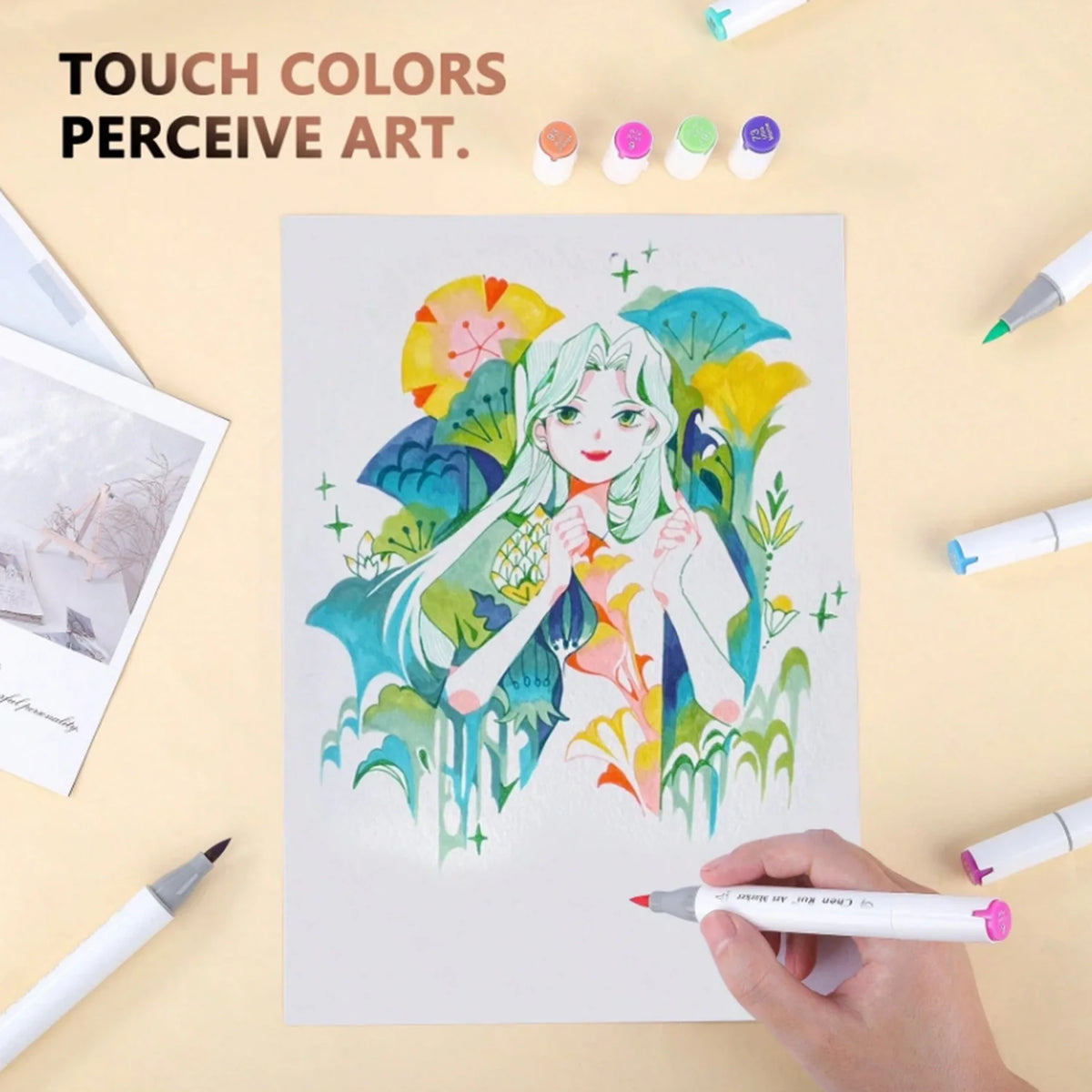 A hand holds an alcohol marker drawing a girl with vibrant colors on white paper surrounded by various art supplies.