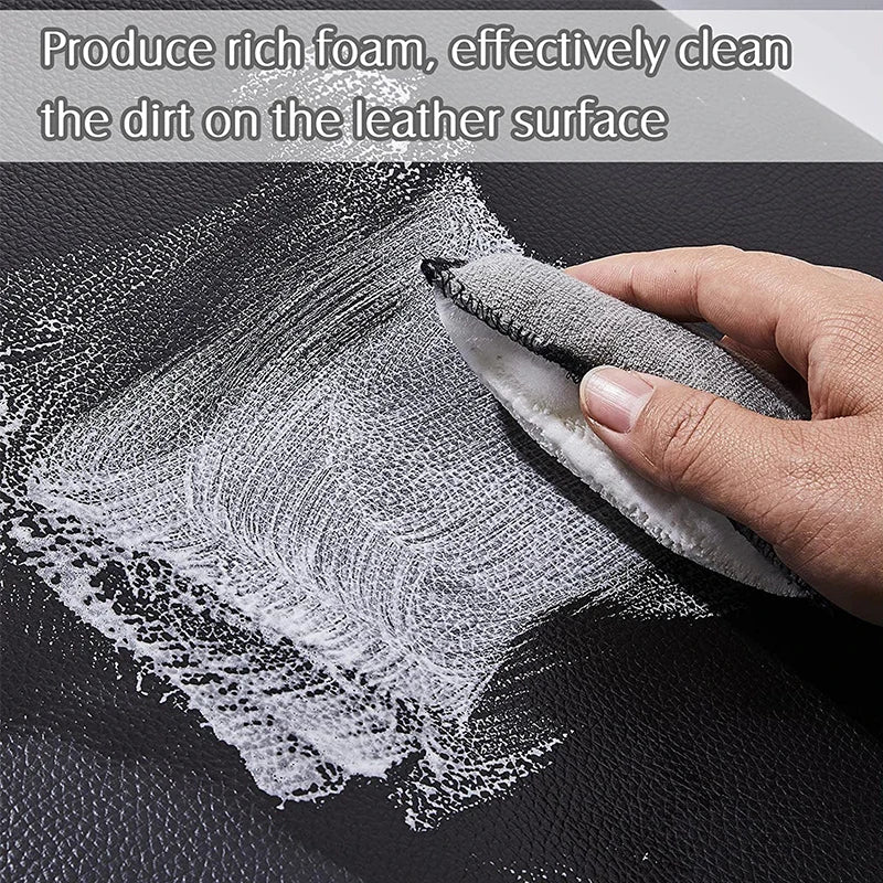 A hand is using a white cleaning sponge to remove dirt from a black leather surface.