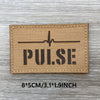 A tan fabric patch with the word PULSE and a heartbeat line printed on it, set against a light wooden background.