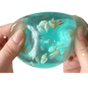 A hand holds a translucent blue liquid containing small seashells and sea creatures, used for sensory play.