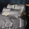 A set of two silky satin pillowcases in gray and off-white are displayed on a bed with dark blue accents.