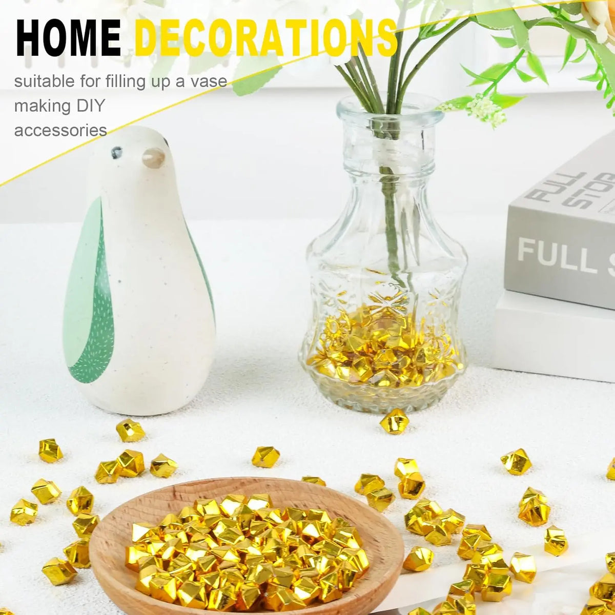 Gold acrylic crushed ice gems scattered on a white surface next to a glass vase and a ceramic bird.