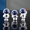 Three handcrafted astronaut figurines in white and blue resin displayed on a reflective surface.
