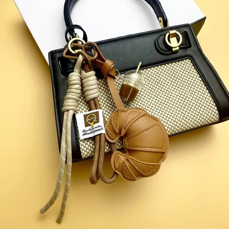 A braided leather croissant keychain with a bag pendant is displayed against a light yellow background.