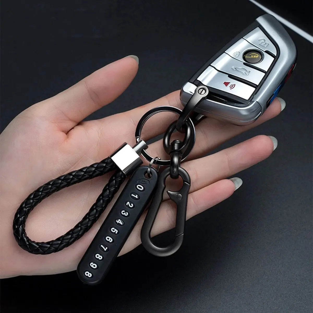 A hand holds a personalized phone number keychain with a silver phone and a braided black leather rope.