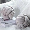A soft gray knitted velvet throw pillow with a tightly knotted design resting on a white textured fabric.