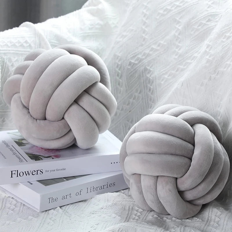 A soft gray knitted velvet throw pillow with a tightly knotted design resting on a white textured fabric.