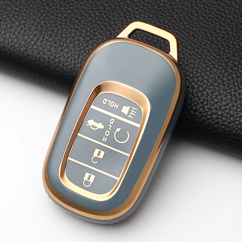A close-up view of a metallic gold Honda key fob cover with buttons and a protective case resting on a black fabric.