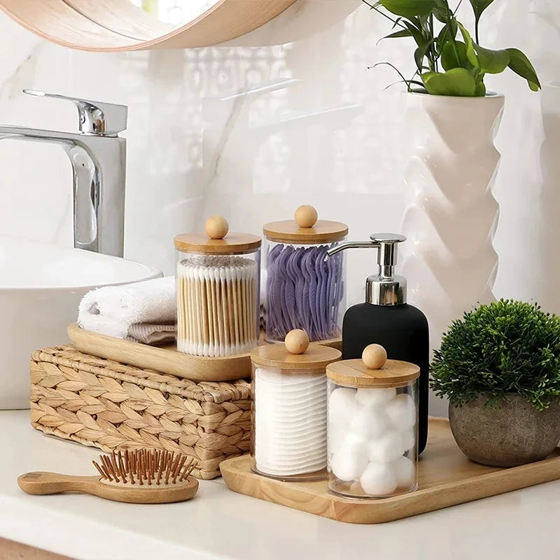 A collection of white bath salts and natural soaps in clear acrylic apothecary jars with wooden lids on a light countertop.