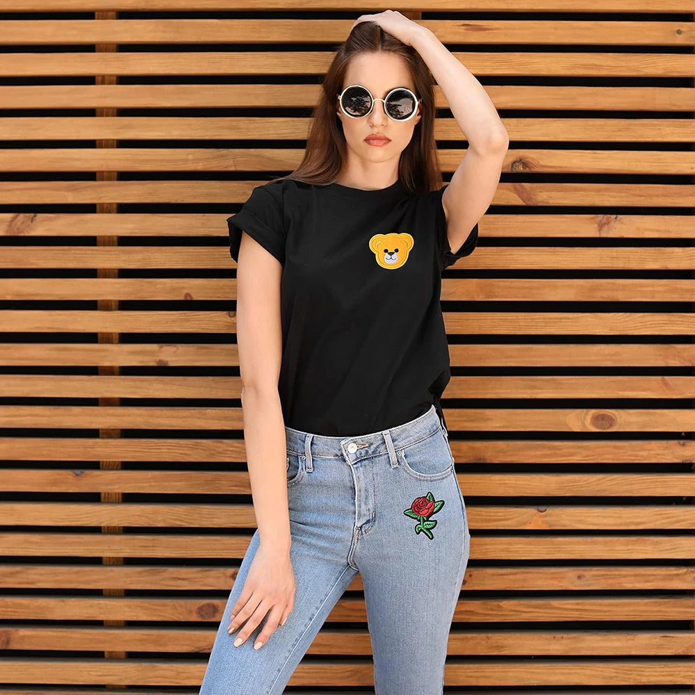A woman models a black t-shirt with a yellow embroidered patch and blue jeans against a wooden background.