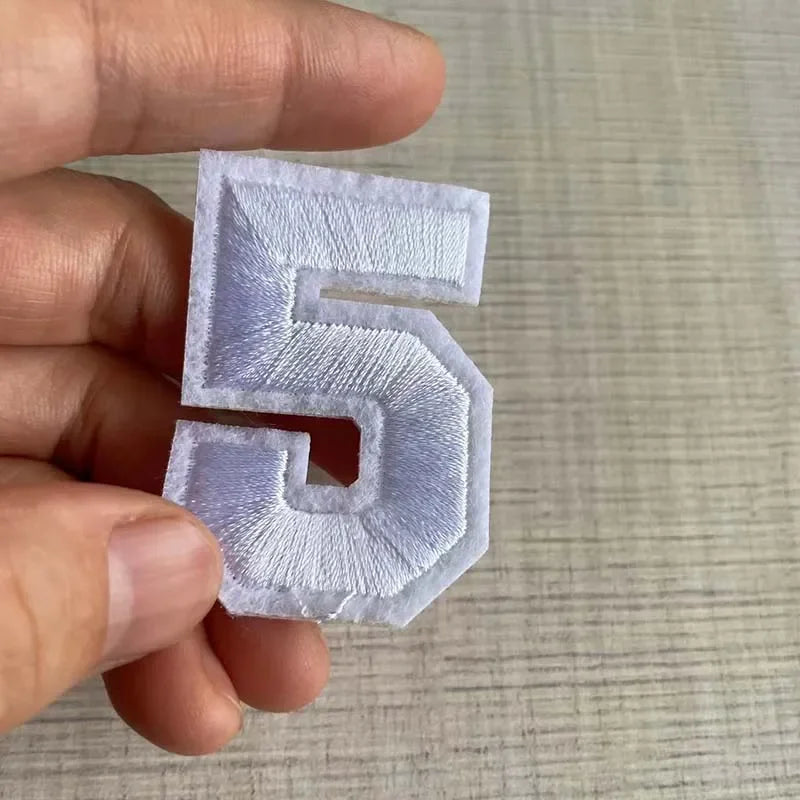 A hand holds a white iron-on 3D number patch shaped like the numeral five.