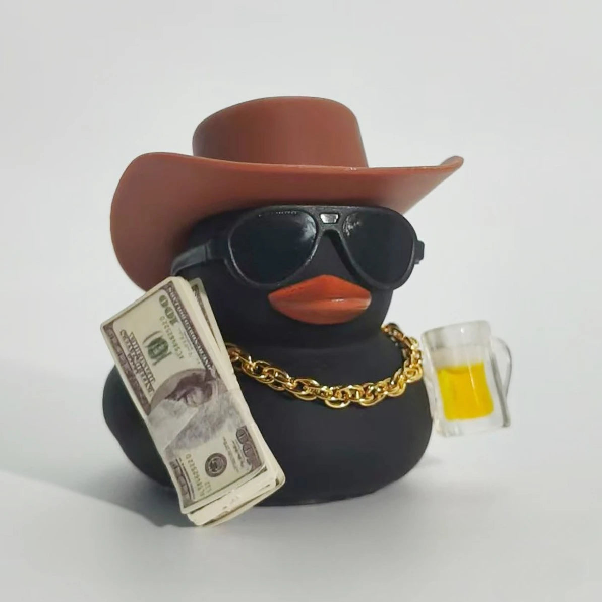 A black rubber duck with a brown cowboy hat, sunglasses, and a gold chain, next to a stack of money and a glass of yellow liq