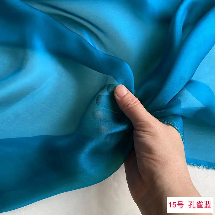 A hand is folding a piece of bright blue silk chiffon fabric to show its drape and texture.