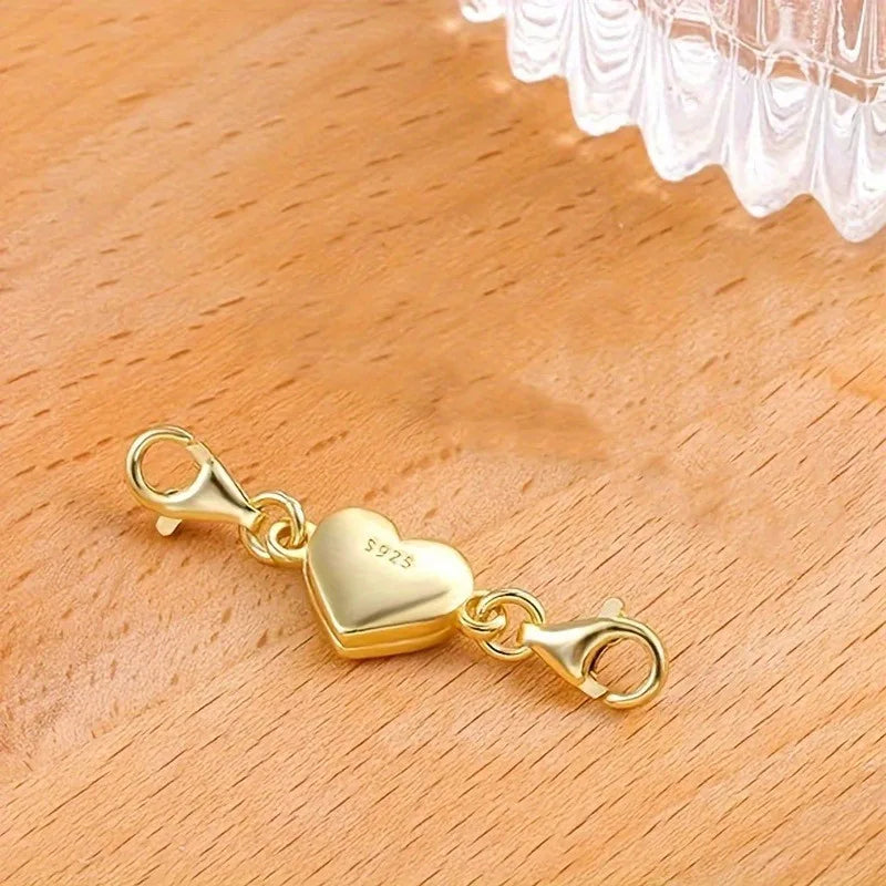 A gold heart-shaped magnetic clasp for necklaces and bracelets resting on a wooden surface.