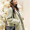 A woman wearing a tweed coat and knit hat holds a black compact square crossbody bag in a festive holiday setting.