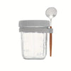 A clear glass overnight oats cup with a lid and a wooden spoon, used for portable breakfast.